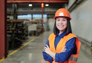 Portrait of smiling confident young Asian factory engineer in orange hardhat and vest standing in workshop