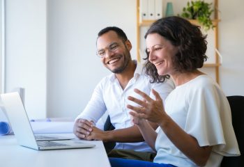 Cheerful colleagues using laptop for video call. Man and woman sitting at workplace together, looking at monitor and laughing. Communication concept