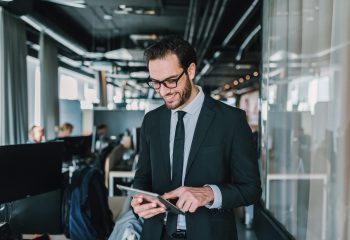 Close up of businessman using tablet while standing in the office. In background coworkers working on computers.