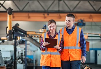 Male and Female Industrial worker use tablet and Walking Through Heavy Industry Manufacturing Factory. They Wear Hard Hats and Safety Jacket