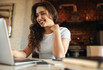 Joyful young female having break while working from home, sitting at desk in front of open laptop, smiling, chatting online via video conference call with friends. Electronic gadgets and communication