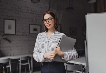 Portrait of brown-eyed business woman in black and white outfit and stylish glasses posing with laptop in white room